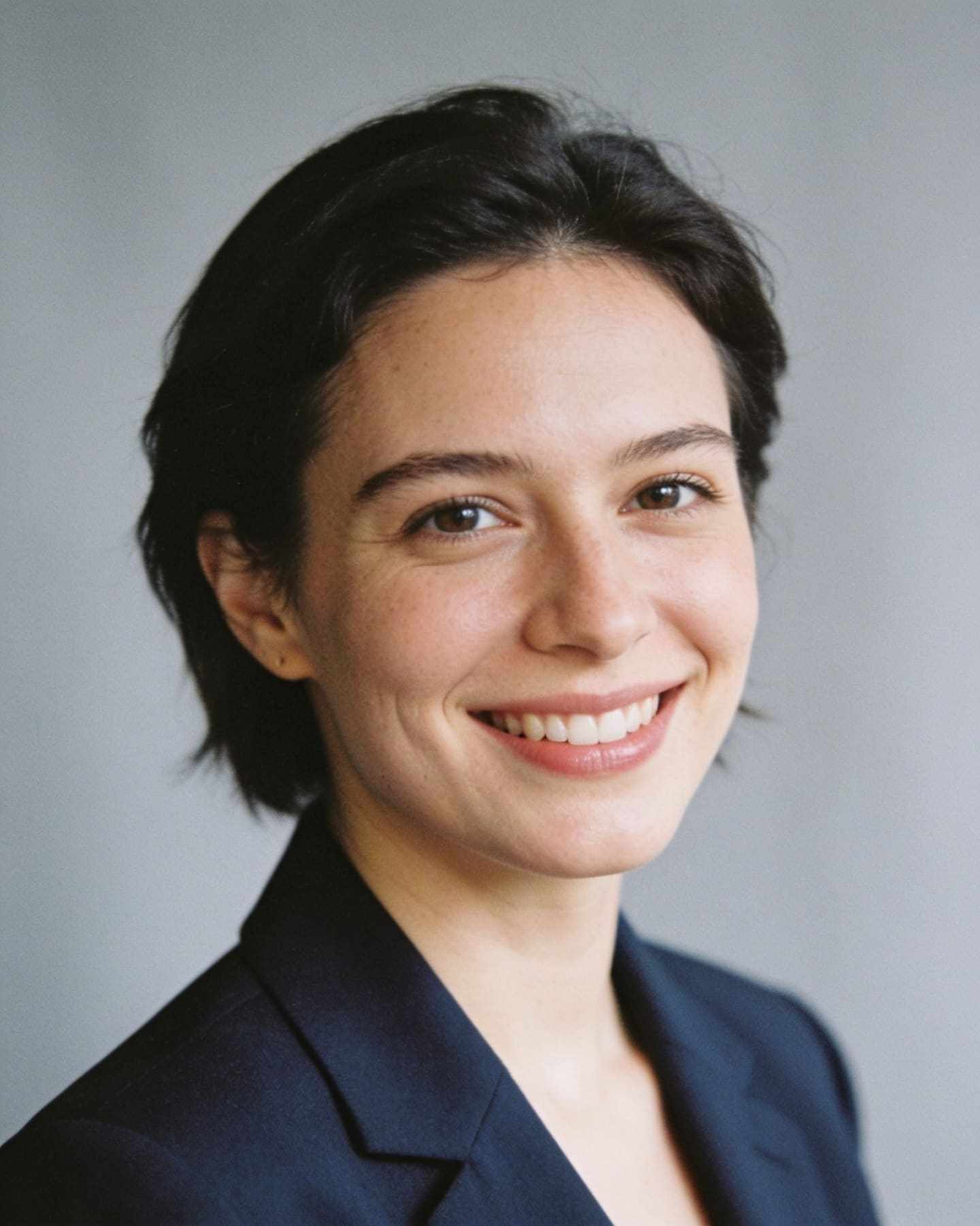 A professional headshot of a smiling woman in a navy blazer against a light grey backdrop.