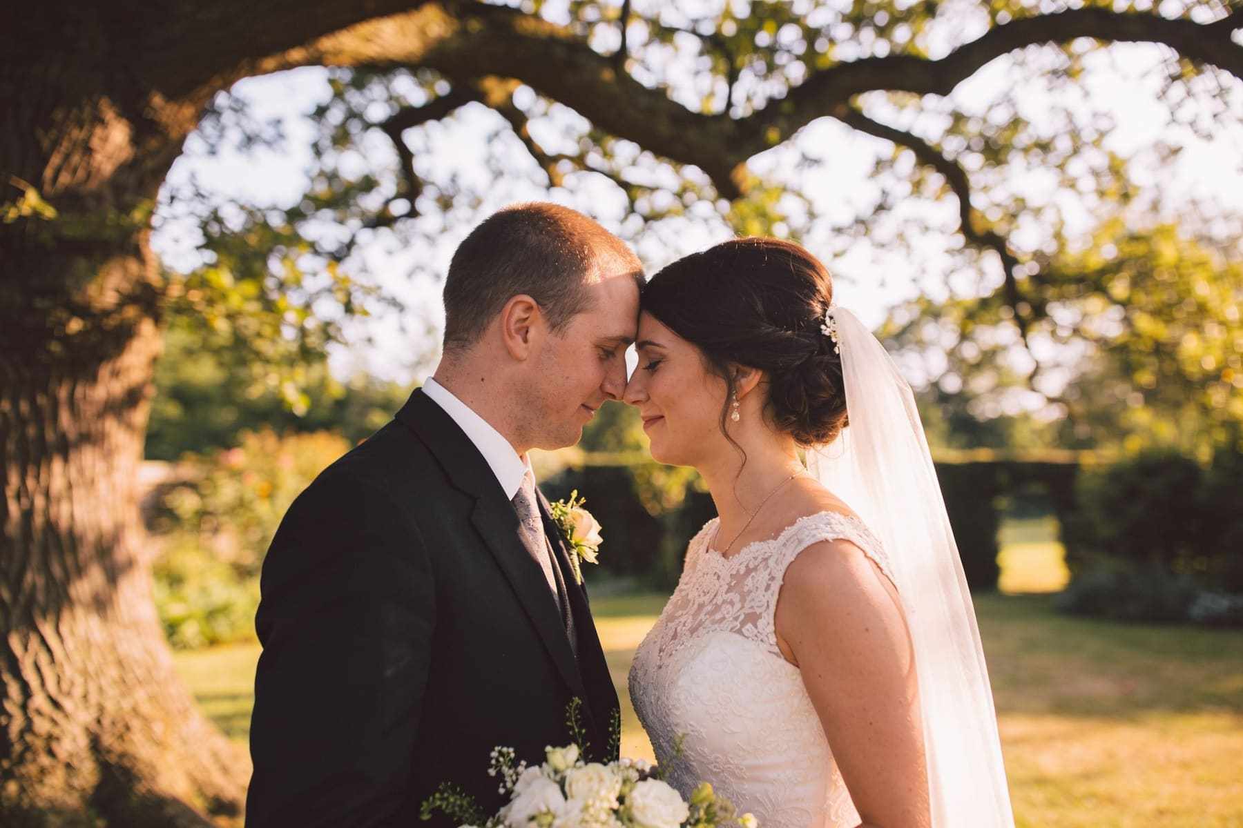 A bride and groom resting their foreheads together under an old oak tree in late afternoon light.