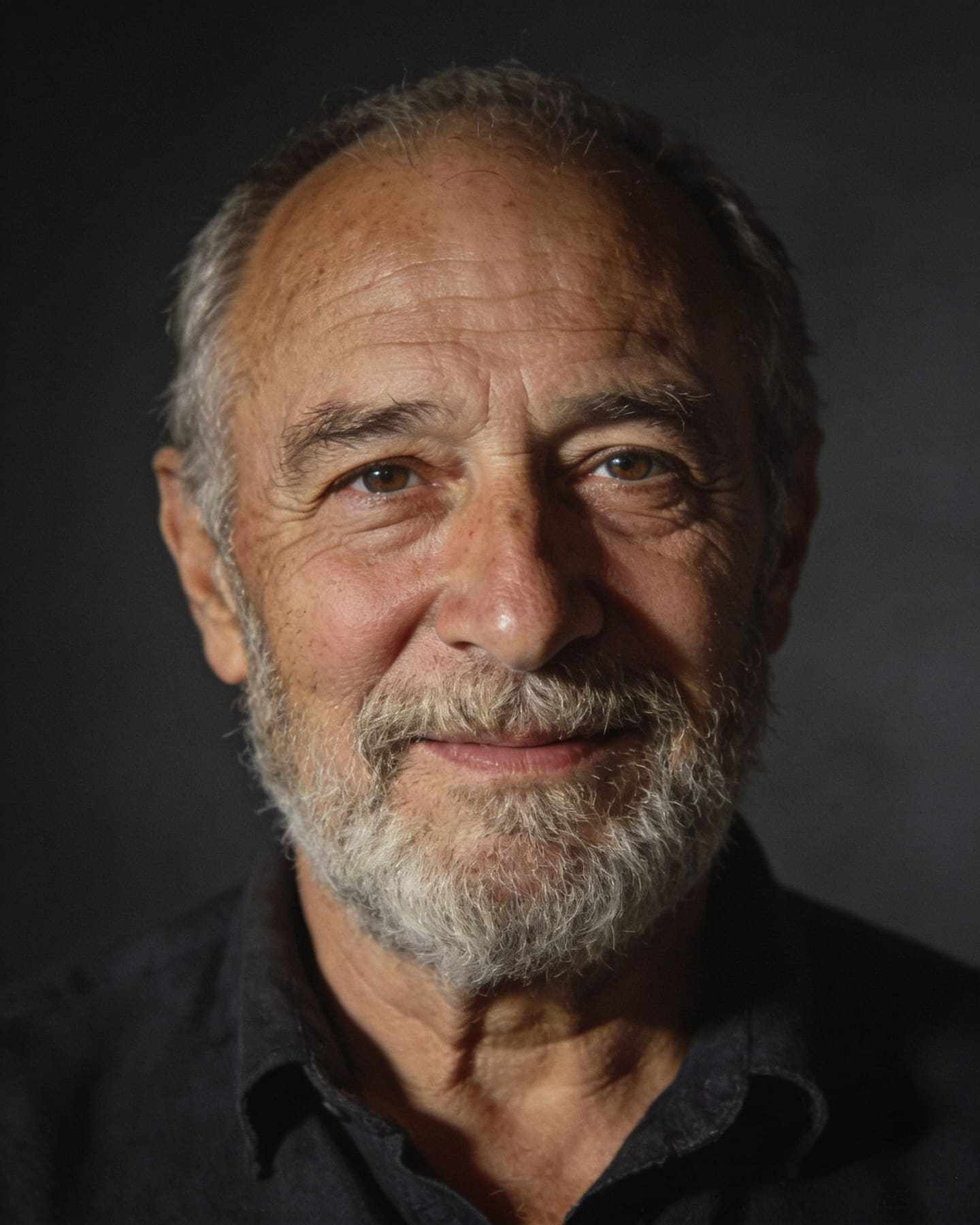 A studio portrait of a man in his sixties with a grey beard and kind eyes, lit from the side.