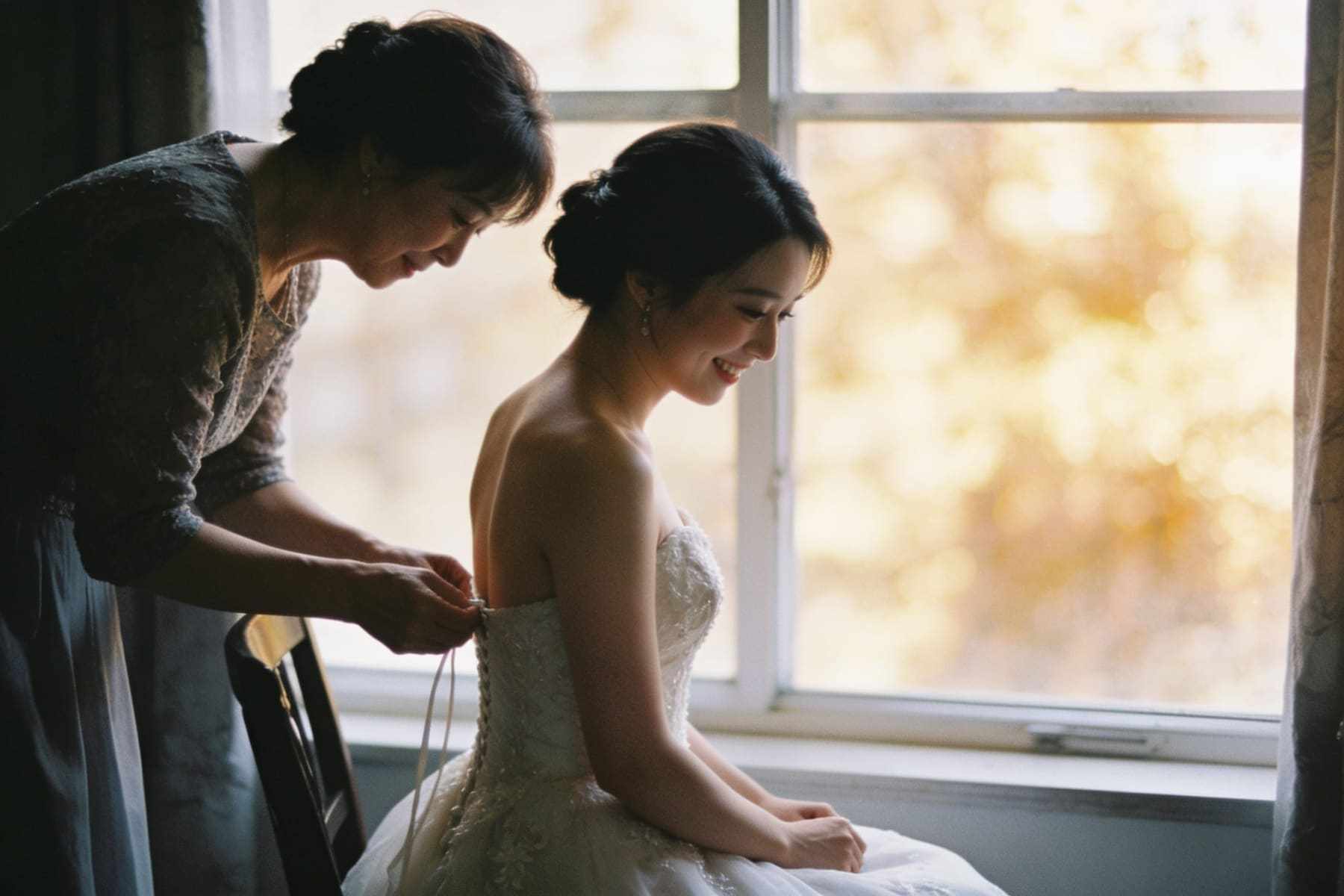 The bride's mother fastening her dress by a window.