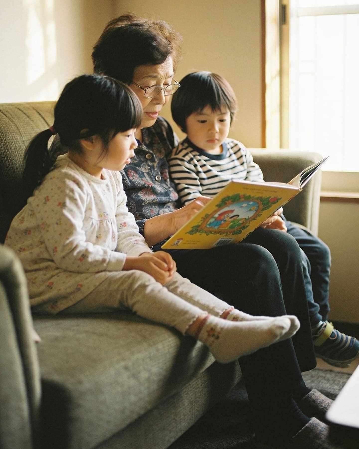A grandmother reading a picture book to two grandchildren on a sofa.