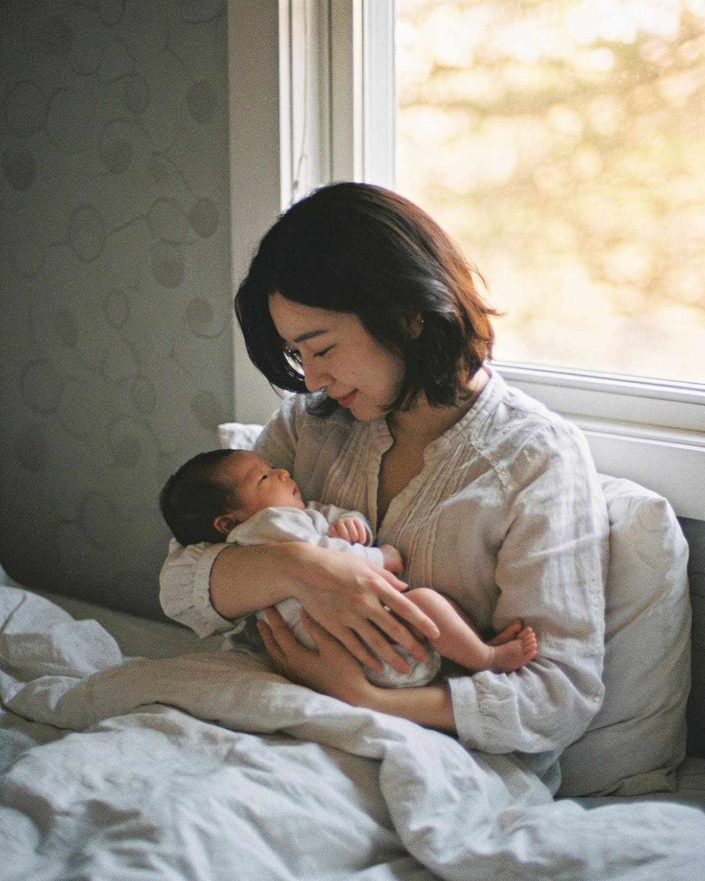A mother cradling her newborn on a bed by a window.