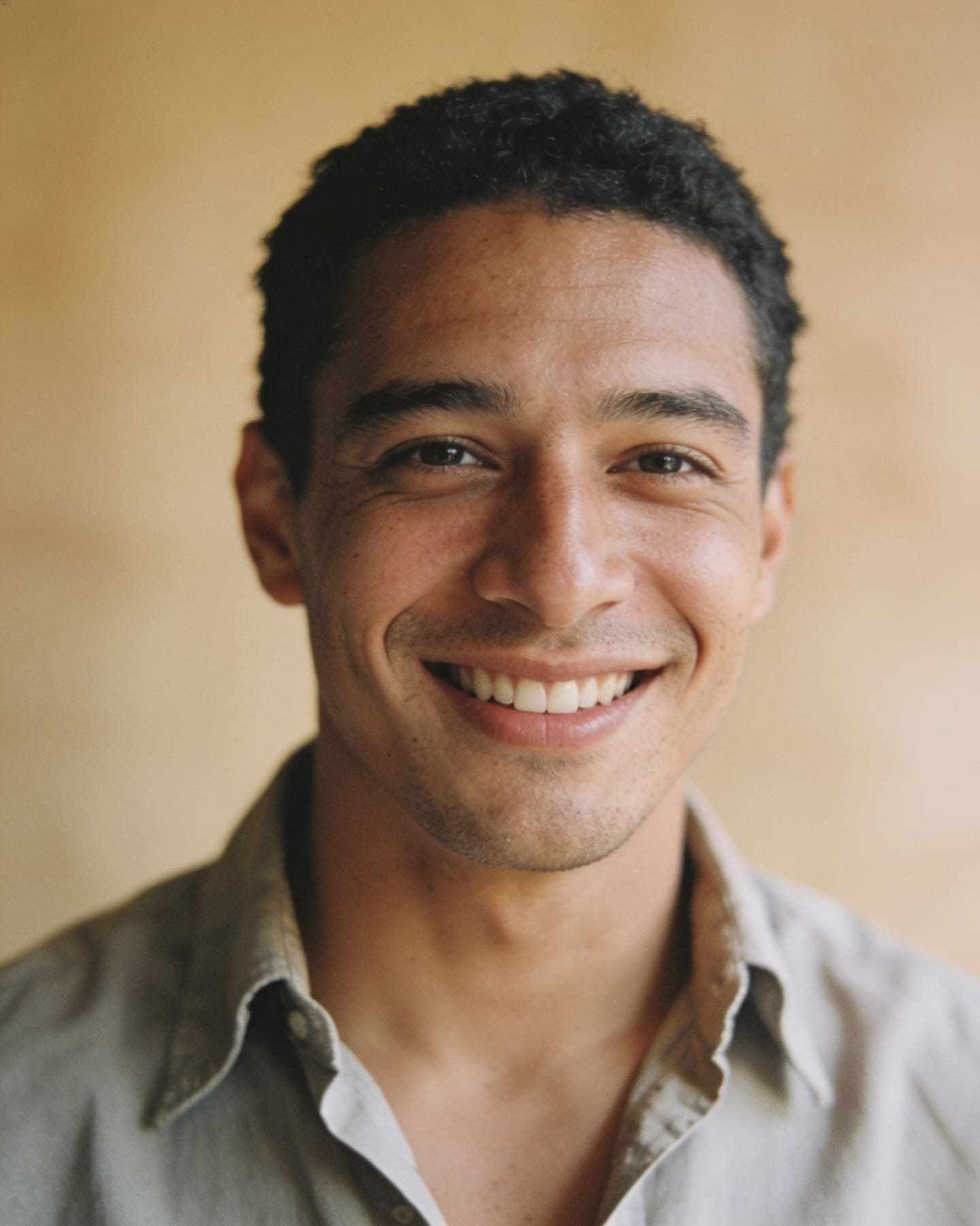 A headshot of a smiling man in an open-collar shirt on a warm beige backdrop.