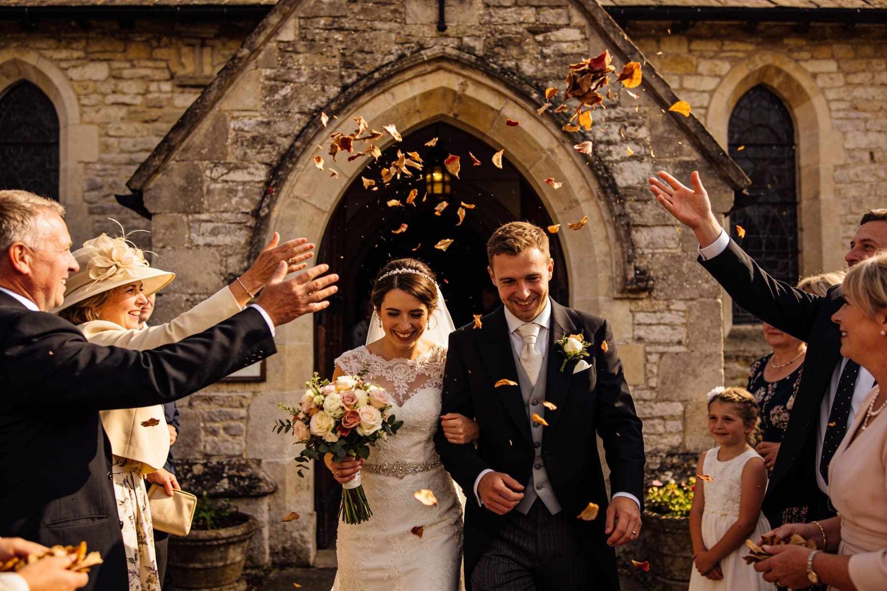 Guests throwing petals as the couple walk out of a stone village church.