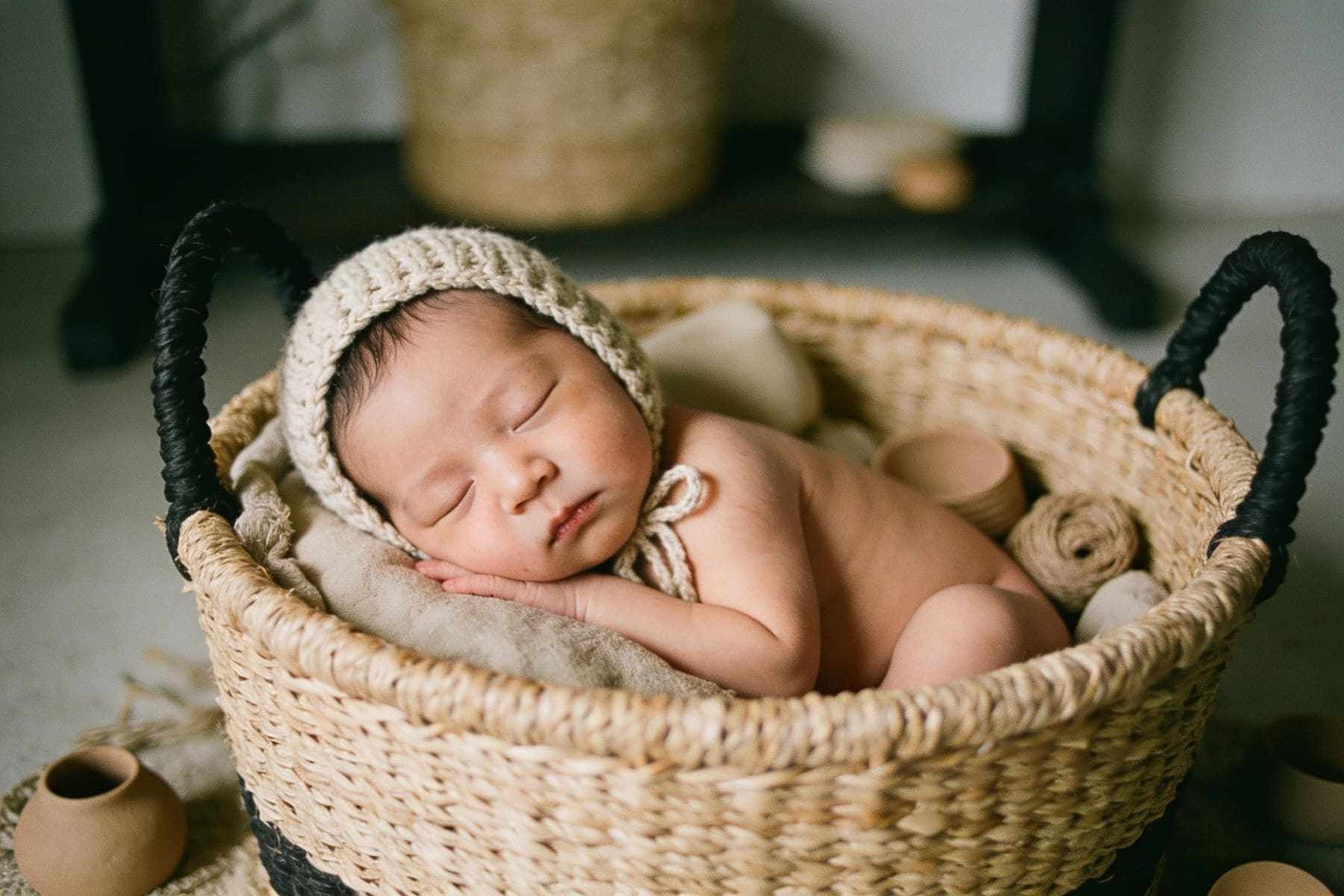 A sleeping newborn in a woven basket wearing a knitted bonnet.