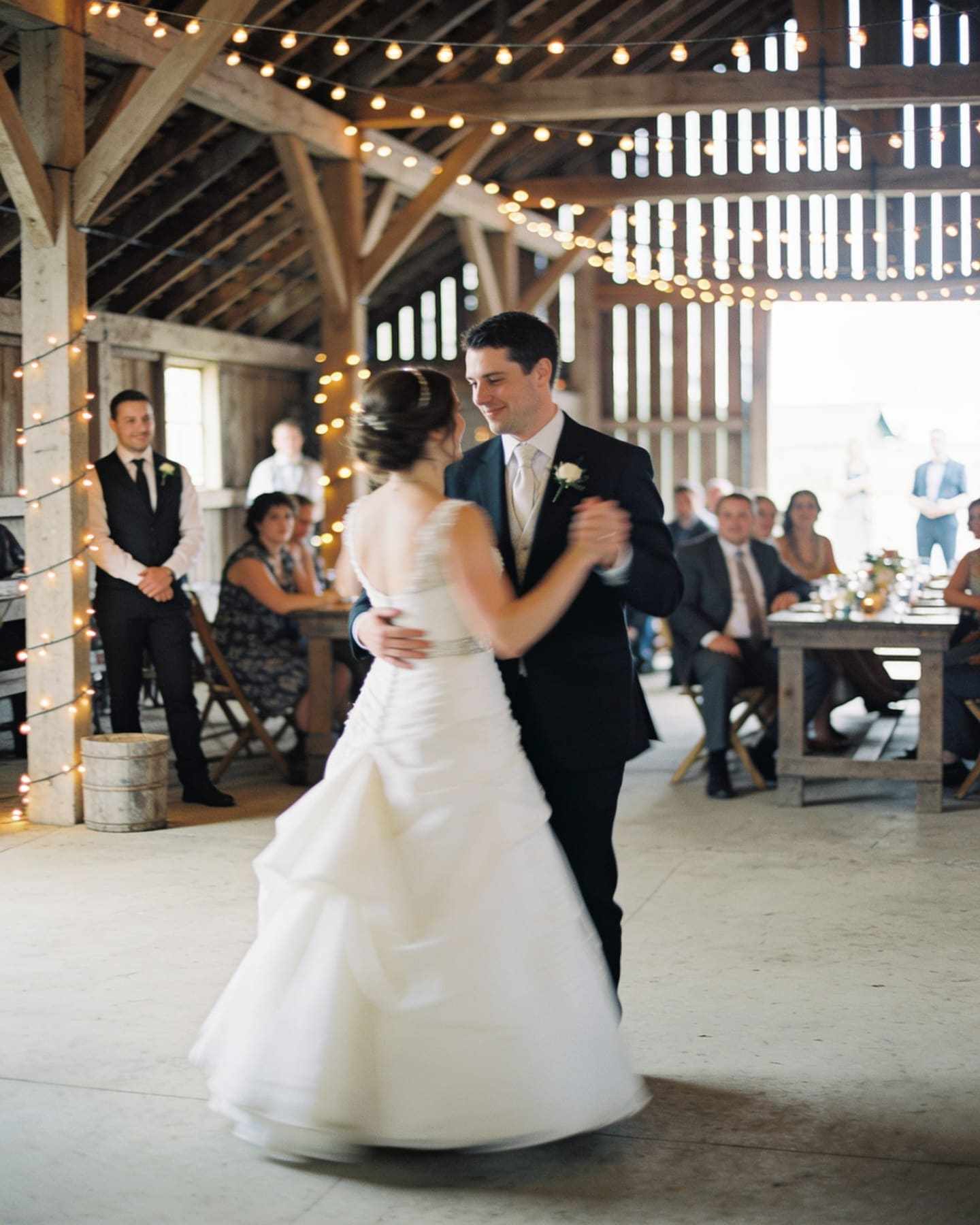 A couple's first dance in a barn lit by festoon lights.